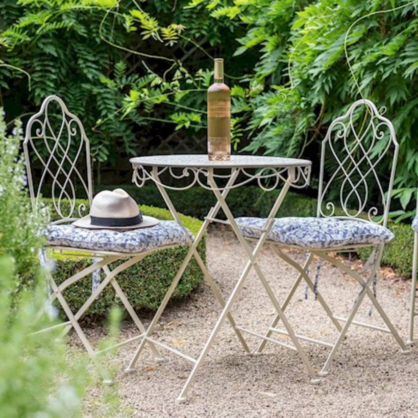 Cream Avalon 3-piece bistro set on a gravel path, with a bottle of wine on the table, blue and white cushions on the chairs, and a boater hat resting on the left chair. Wisteria and box hedges in the background.