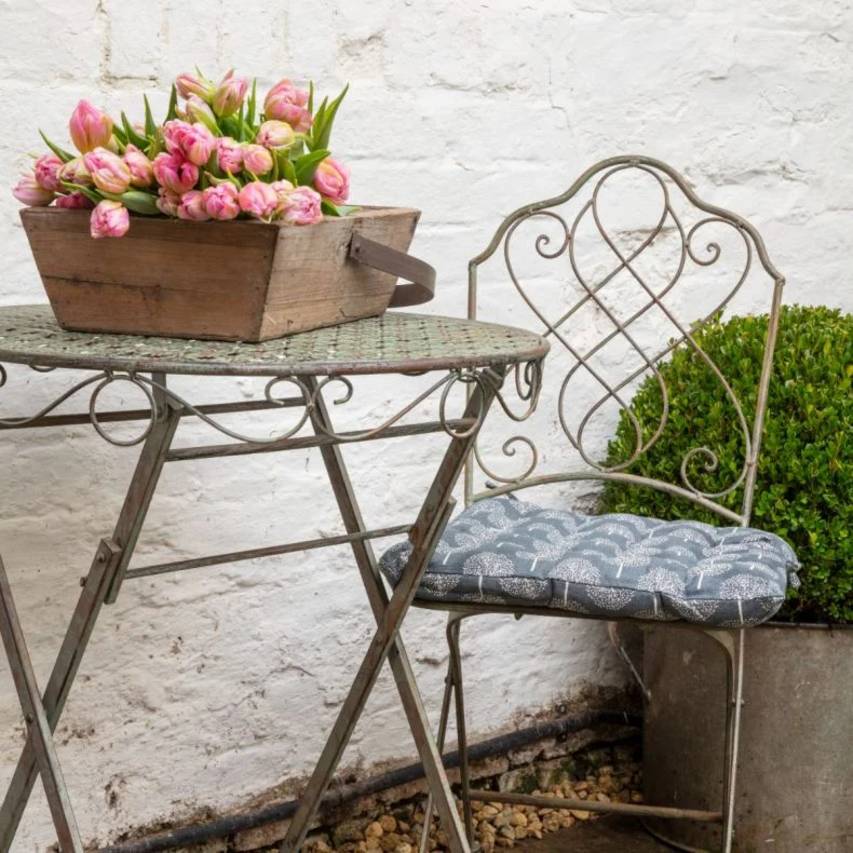 Green rust Avalon bistro chair with a blue cushion and matching table, featuring a wooden trug filled with cut pink tulips. A box ball in a metal planter sits behind the chair.