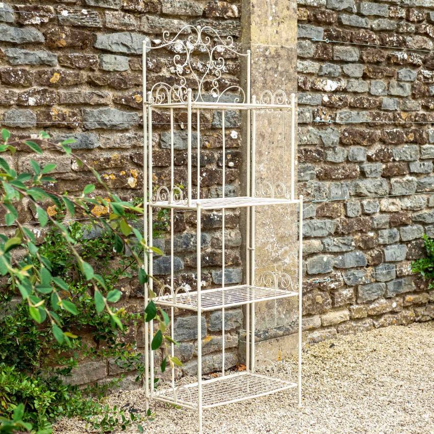 Cream Blaisdon bakers rack with four-tier shelving, placed against a stone wall with plants.
