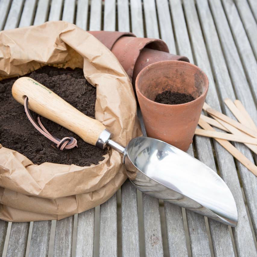 Compost scoop on wooden bench beside terracotta pots and paper bag filled with compost