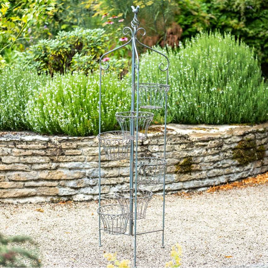 Folding 6 pot holder in antique blue on gravel path in front of low stone wall with shrubs in background.