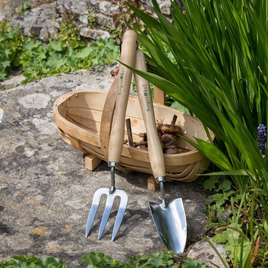 Burgon and Ball RHS mid handled gardening fork and trowel leant against wooden trug resting on lichen-covered stone next to crocosmia
