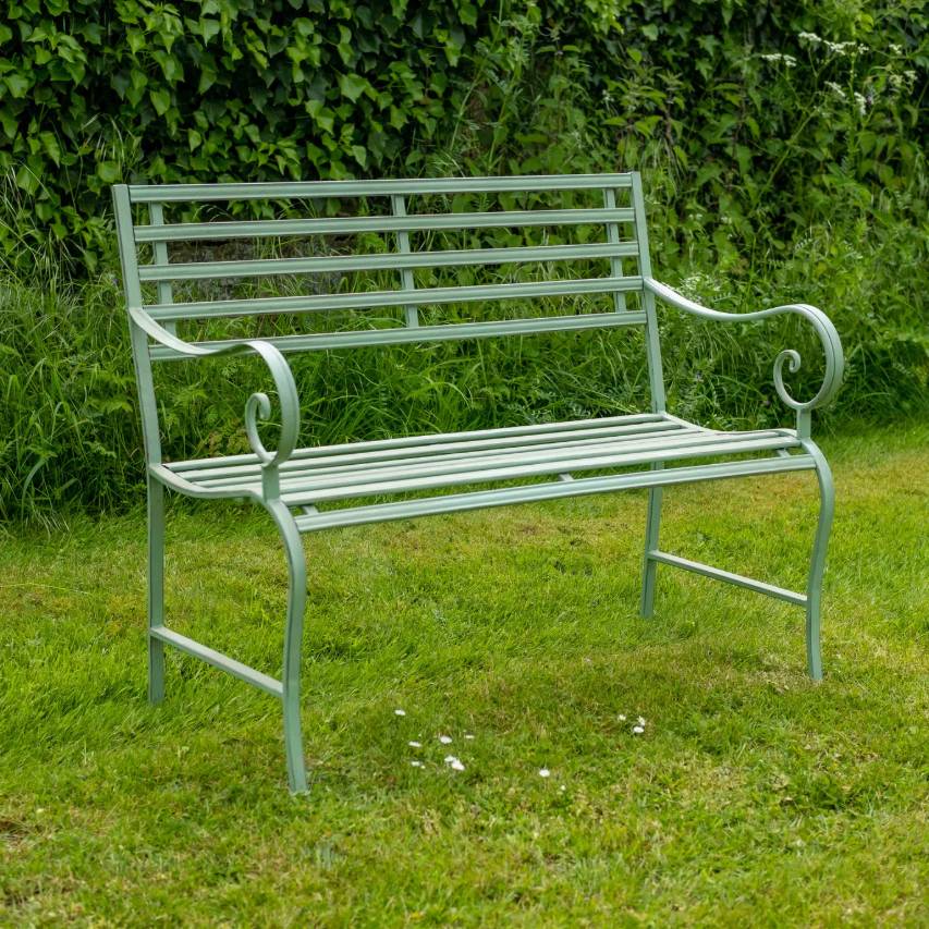 Sage green slatted bench on grass with greenery in the background.