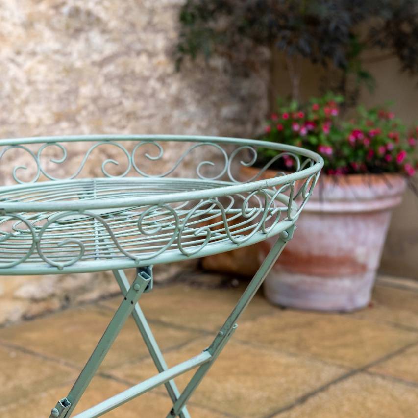 Close-up side view of the green Woodland butler's table on a patio, with a stone wall and a potted fuchsia in the background.